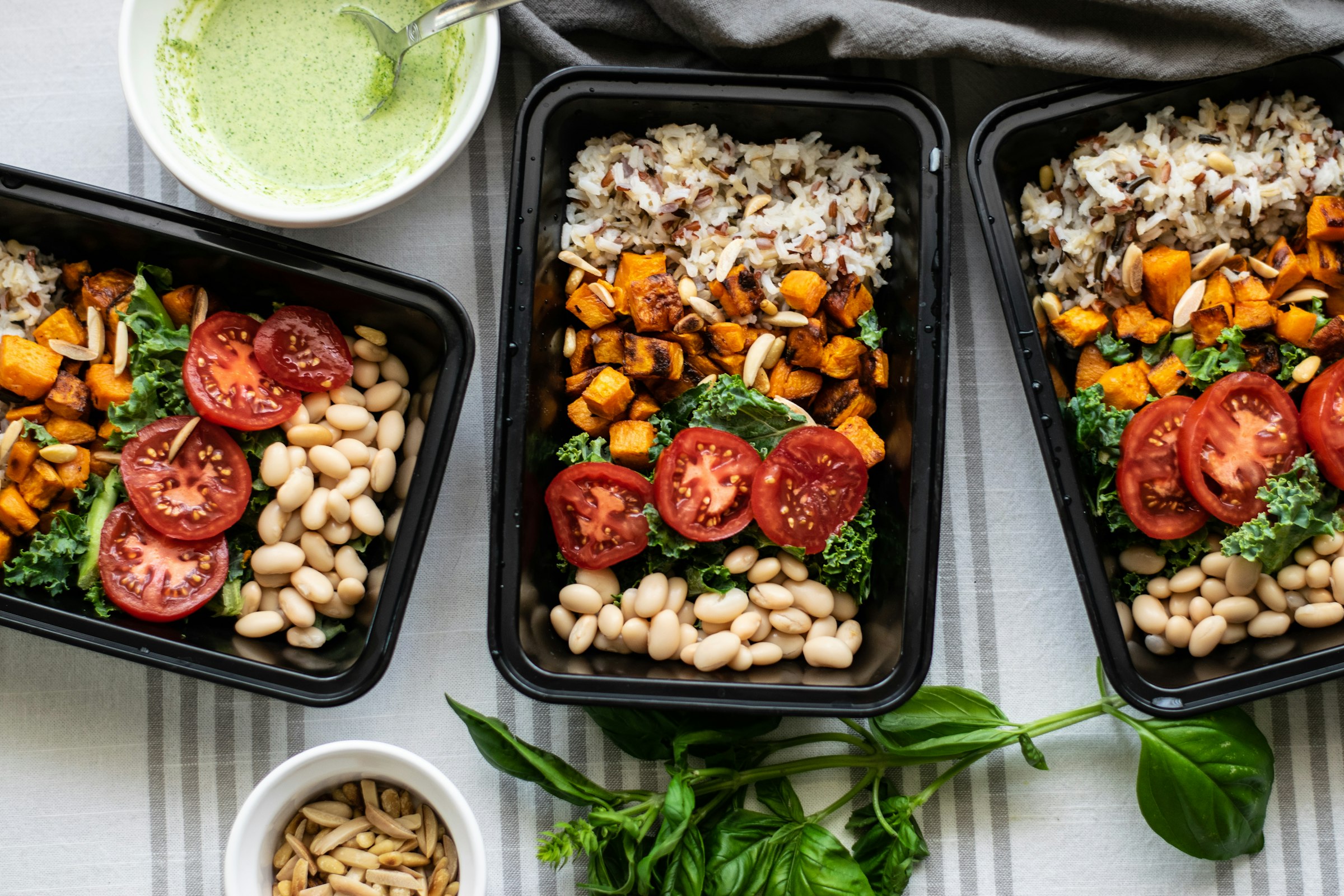 Three black meal prep trays photographed from above, each divided into compartments of protein, grains and greens
